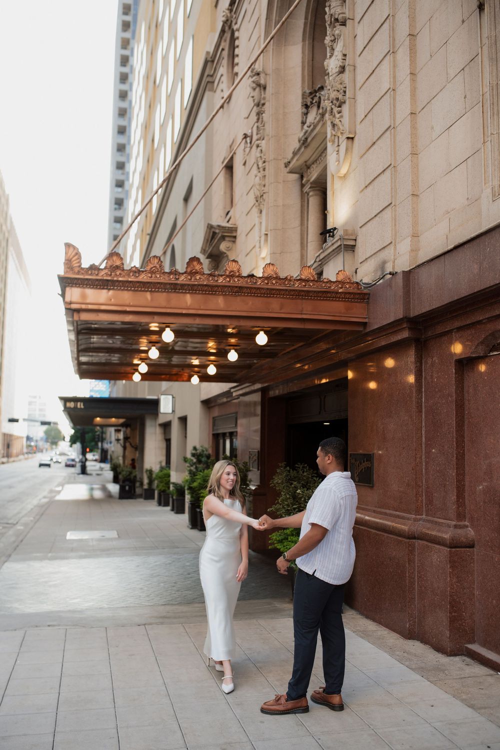 A couple stands together outside an elegant historic building entrance with string lights overhead.