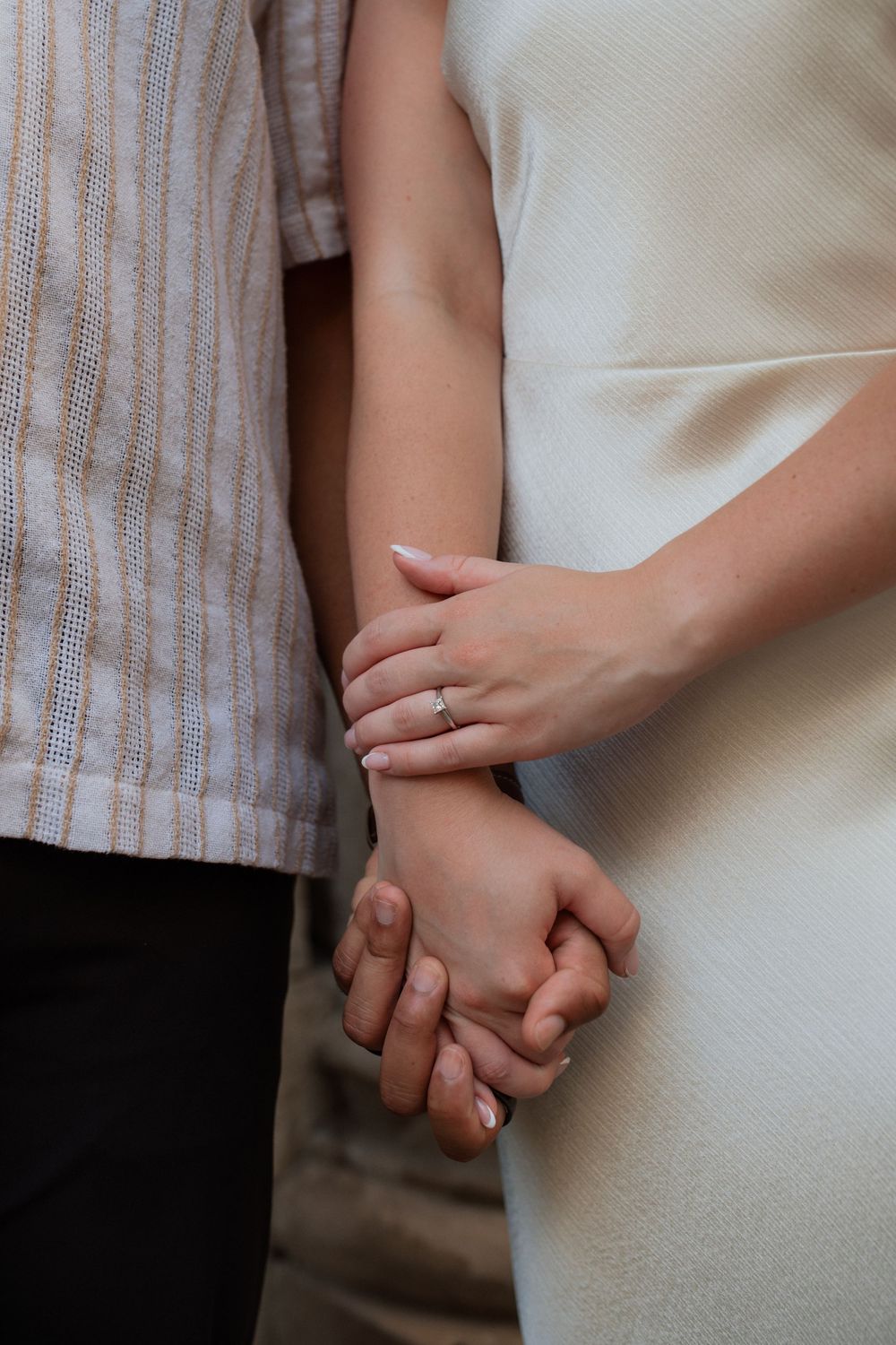 Close-up of couple holding hands tenderly with wedding ring visible, wearing white dress and striped shirt.