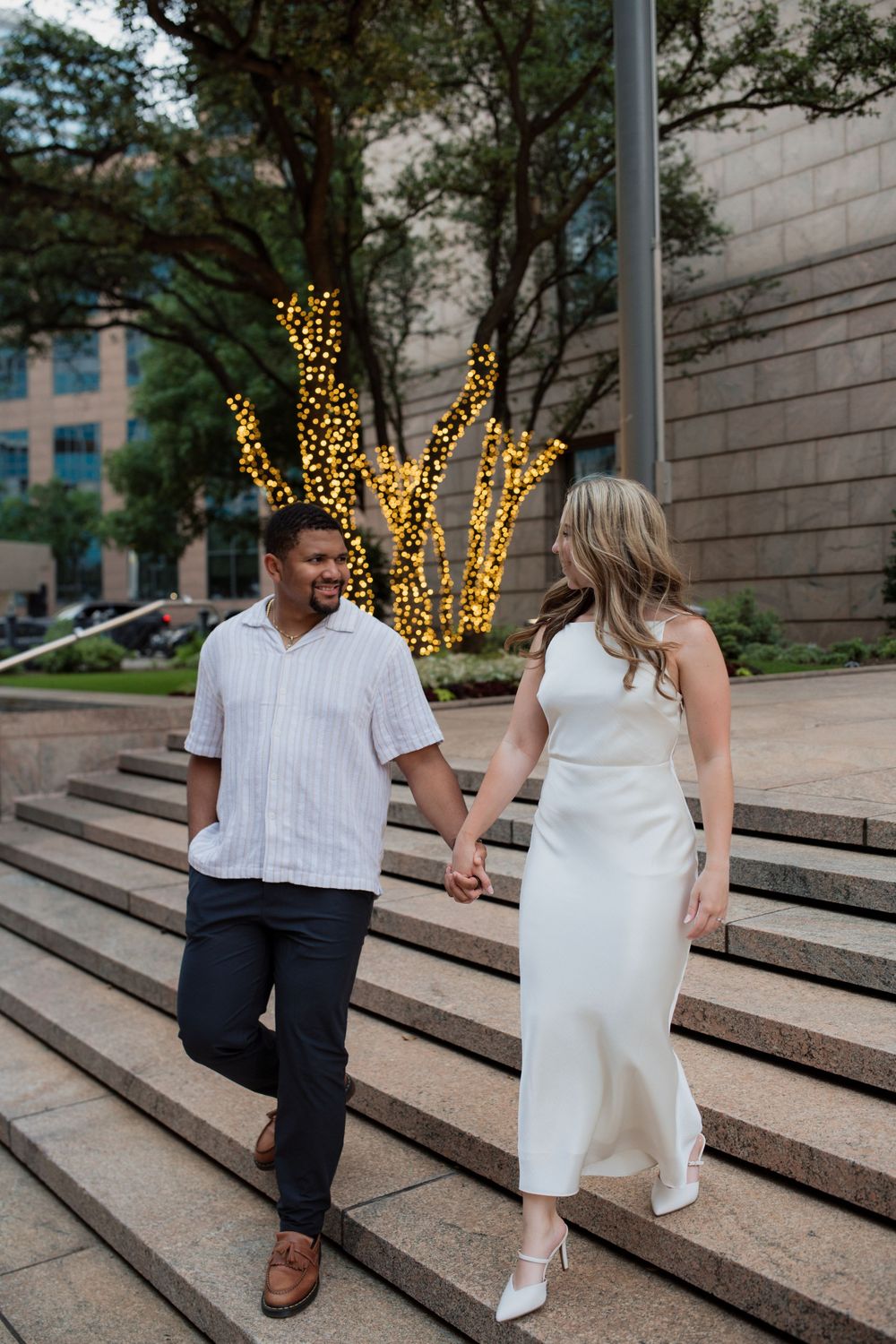 A couple poses on concrete steps with decorative lights in the background during an evening photoshoot.