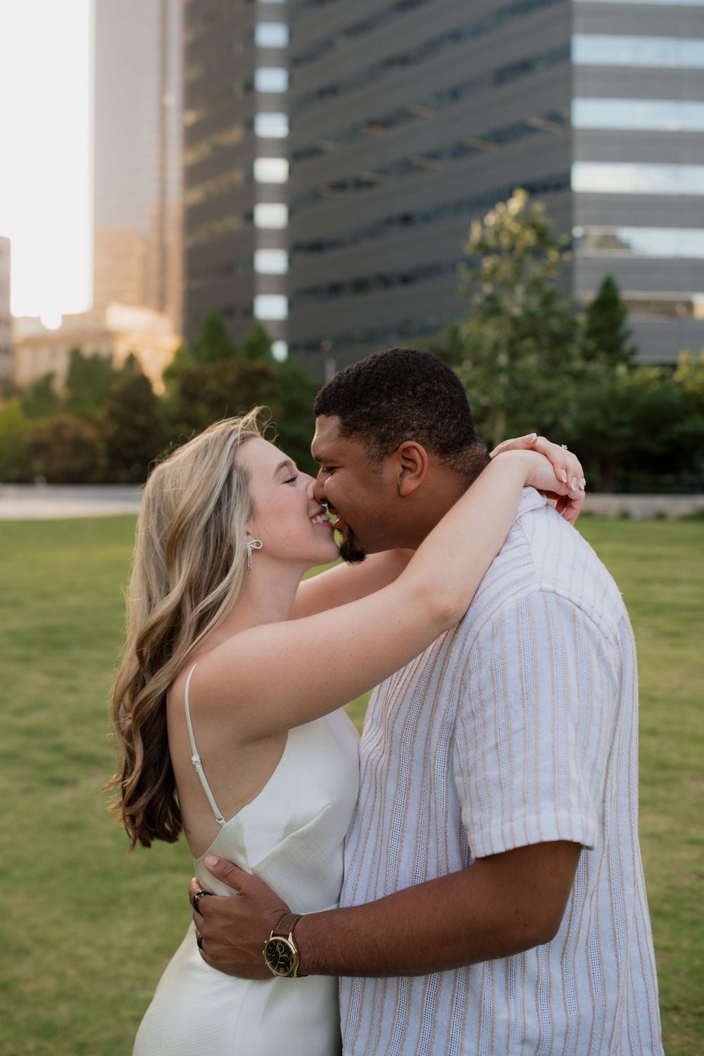 A couple shares an intimate embrace on a grassy lawn with urban buildings in the background at sunset.