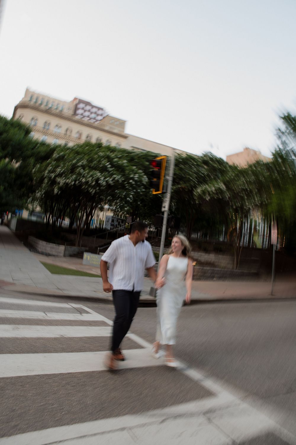 A couple running playfully through a city plaza with modern buildings in the background.