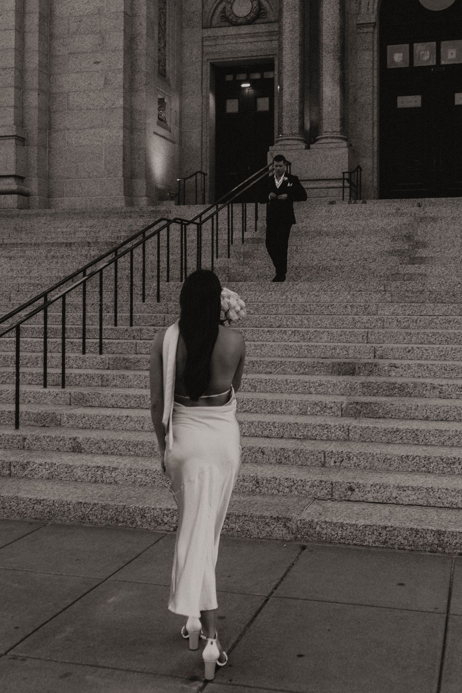 Woman in white dress walking up stone steps of a grand building in a black and white photo.