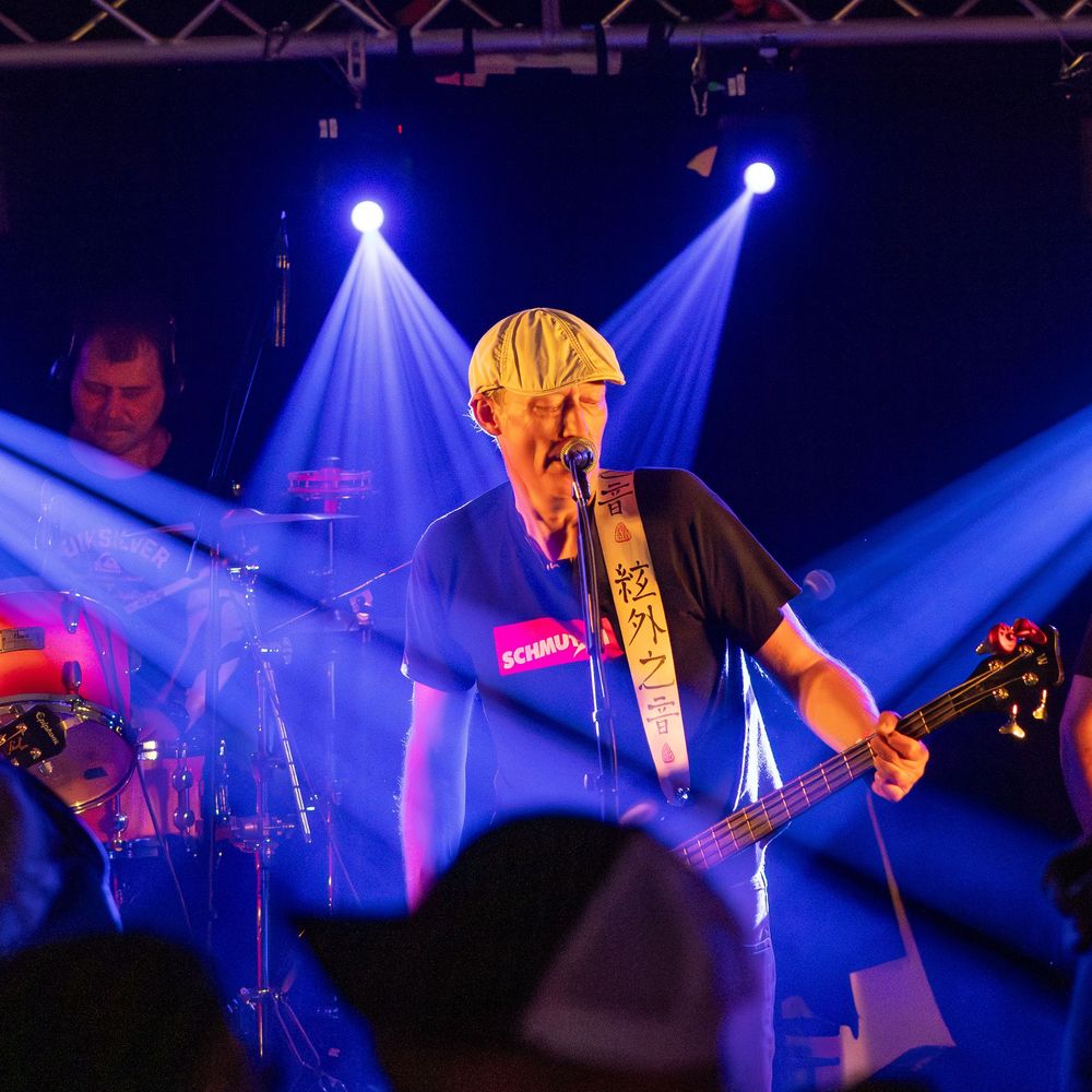 Musicians perform on stage with dramatic blue lighting and fog effects during a rock concert performance.