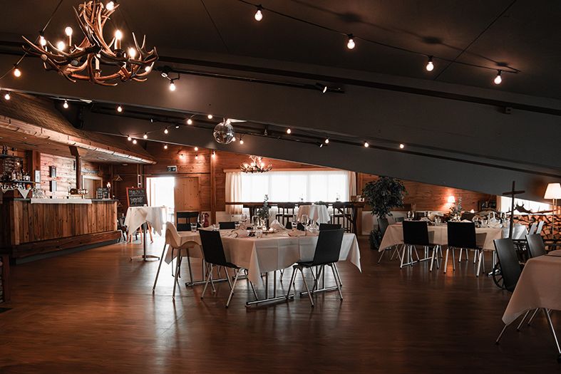 A dimly lit rustic event venue with wooden walls, chandeliers, and scattered tables and chairs creates an intimate atmosphere.