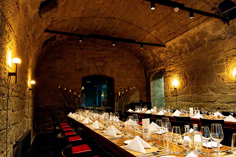 Long elegant dining table set for a formal dinner inside a rustic stone wine cellar with warm lighting and red chairs.