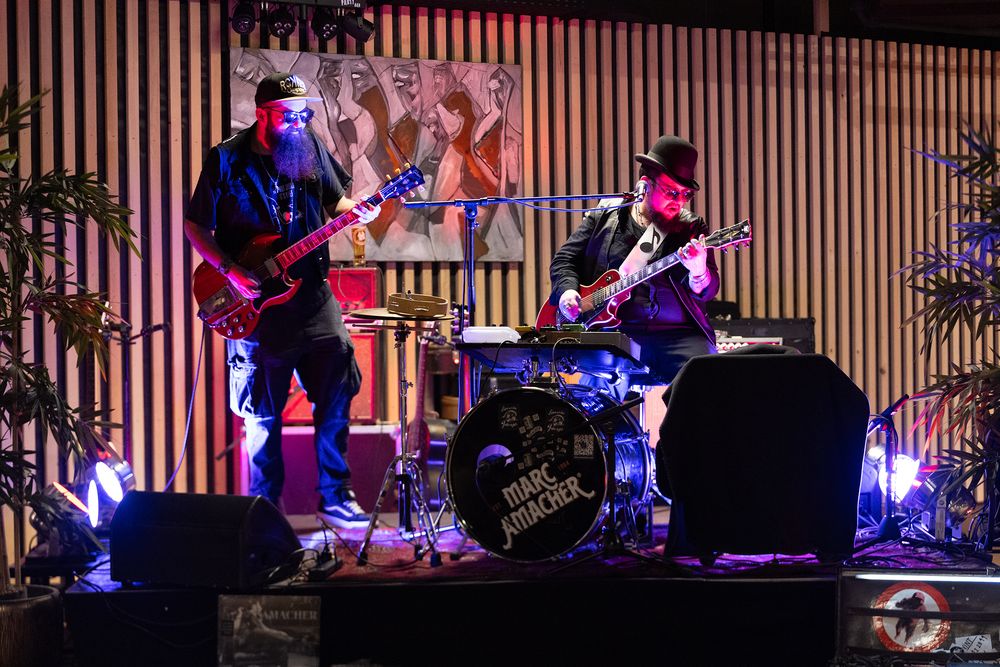 Musicians perform on stage with colorful purple and blue lighting at a live concert venue.