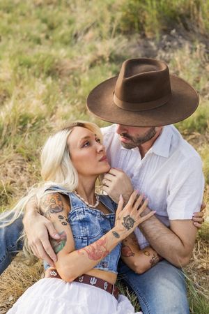 couple sitting in a field, dressed in western style with denim and a brown hat, sharing an intimate moment in grass field in North Carolina