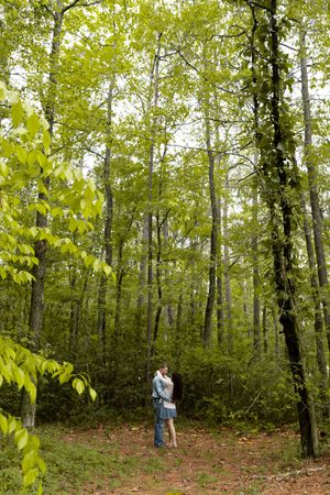 A couple shares a romantic embrace in a lush green forest, surrounded by tall trees and vibrant foliage.