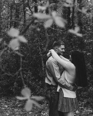 Black and white photo of a couple embracing romantically in an autumn forest, framed by blurred leaves.