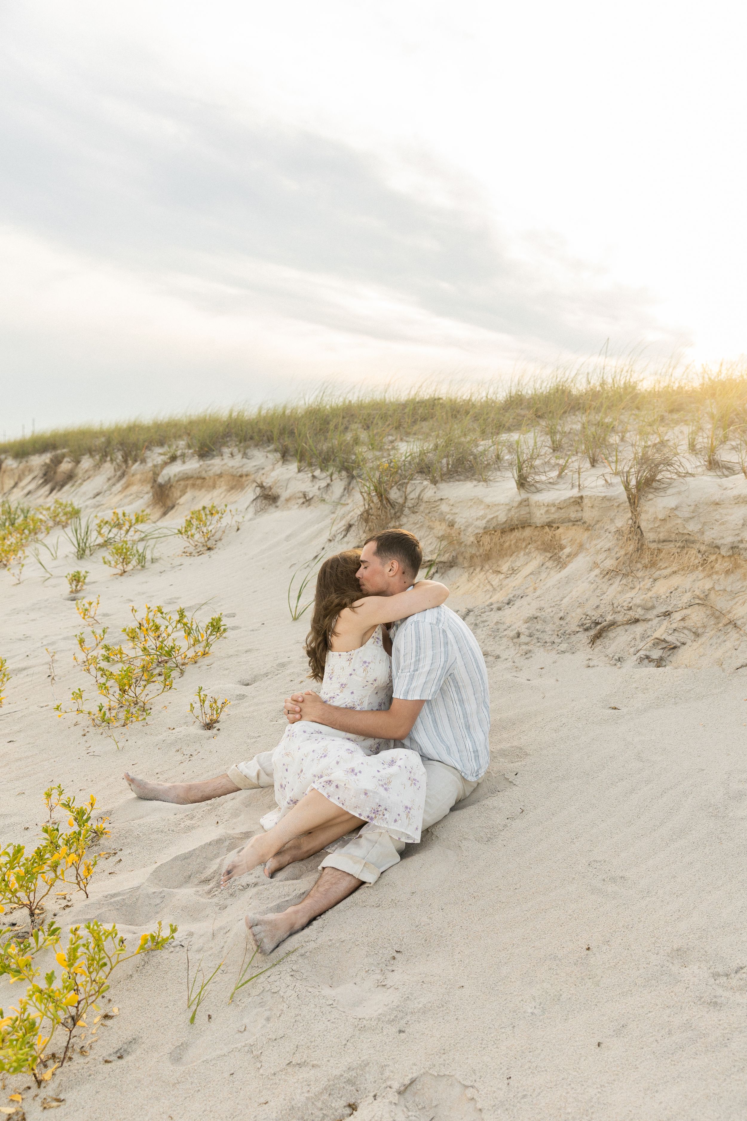 Couple embracing romantically on sandy beach dunes at sunset, woman in floral dress, man in striped shirt.