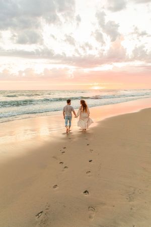 Couple holding hands walking on sandy beach at sunset, leaving footprints, with pastel sky and ocean waves. Costal North Carolina