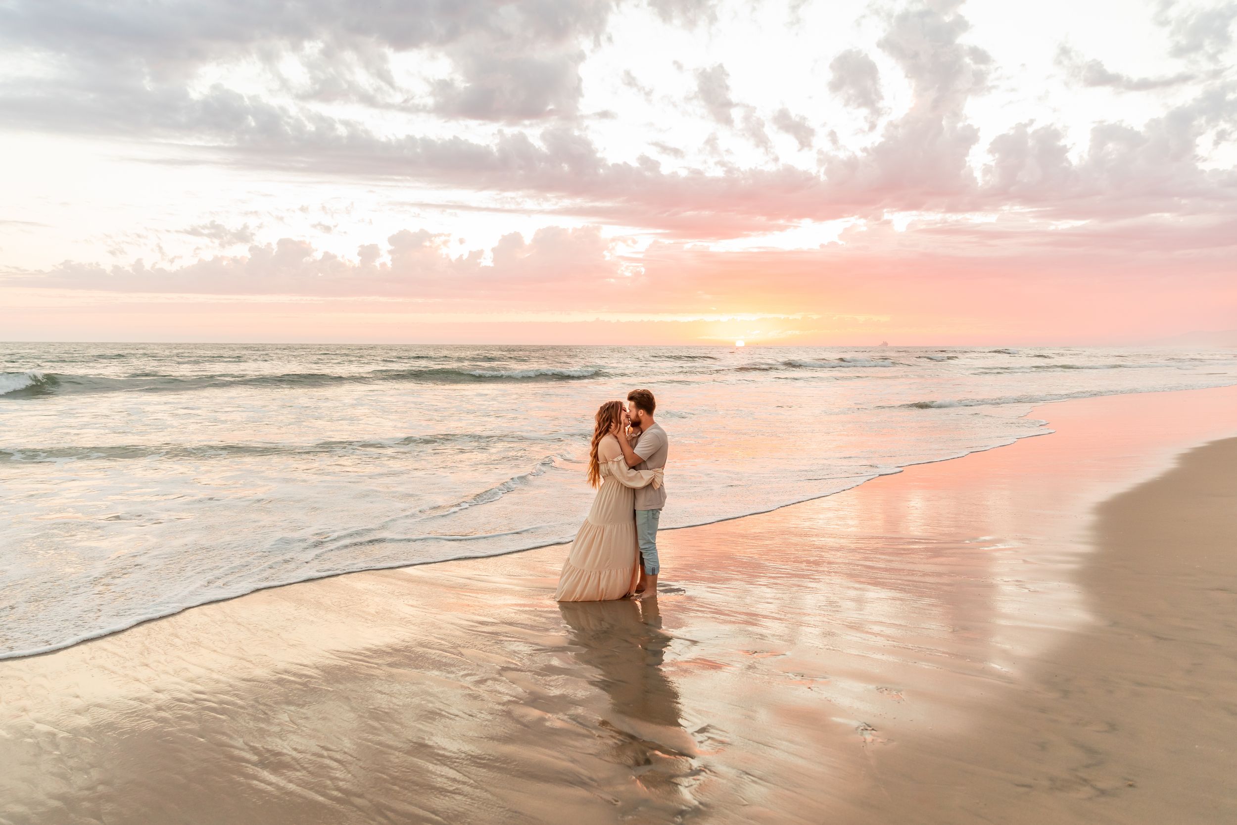 Couple sharing a romantic kiss on a sandy beach o Wilmington North Carolina at sunset, waves gently lapping the shore in soft pink light.