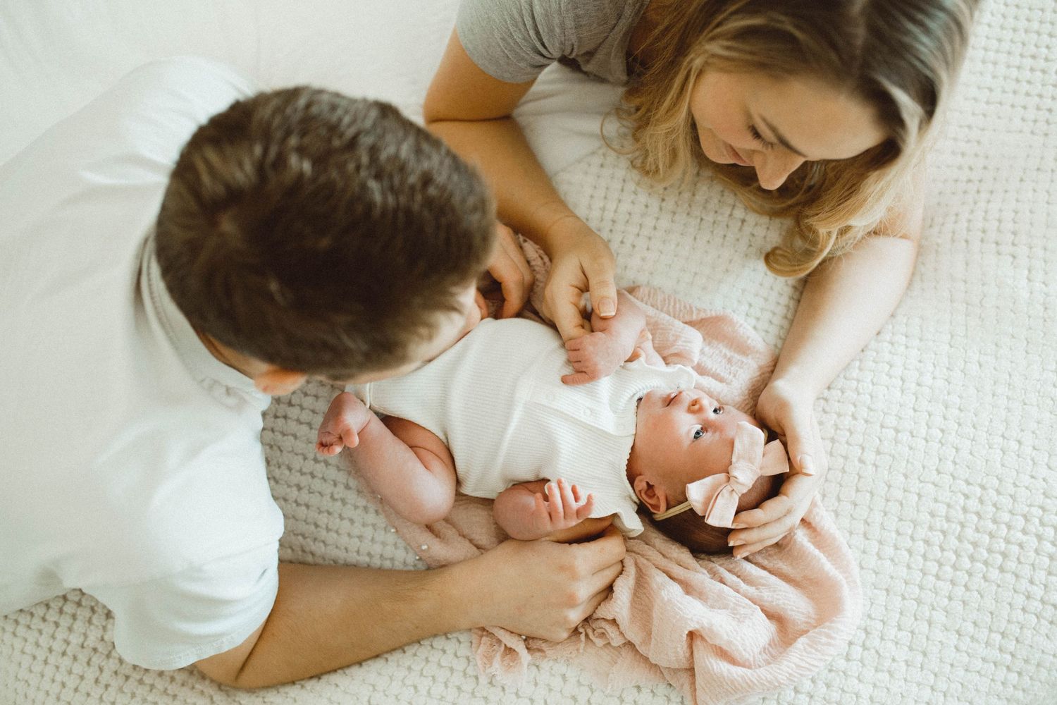 Photo by B.Logsdon in Bluffton, South Carolina of parents lying on white bedding lovingly caring for their newborn baby dressed in pink.