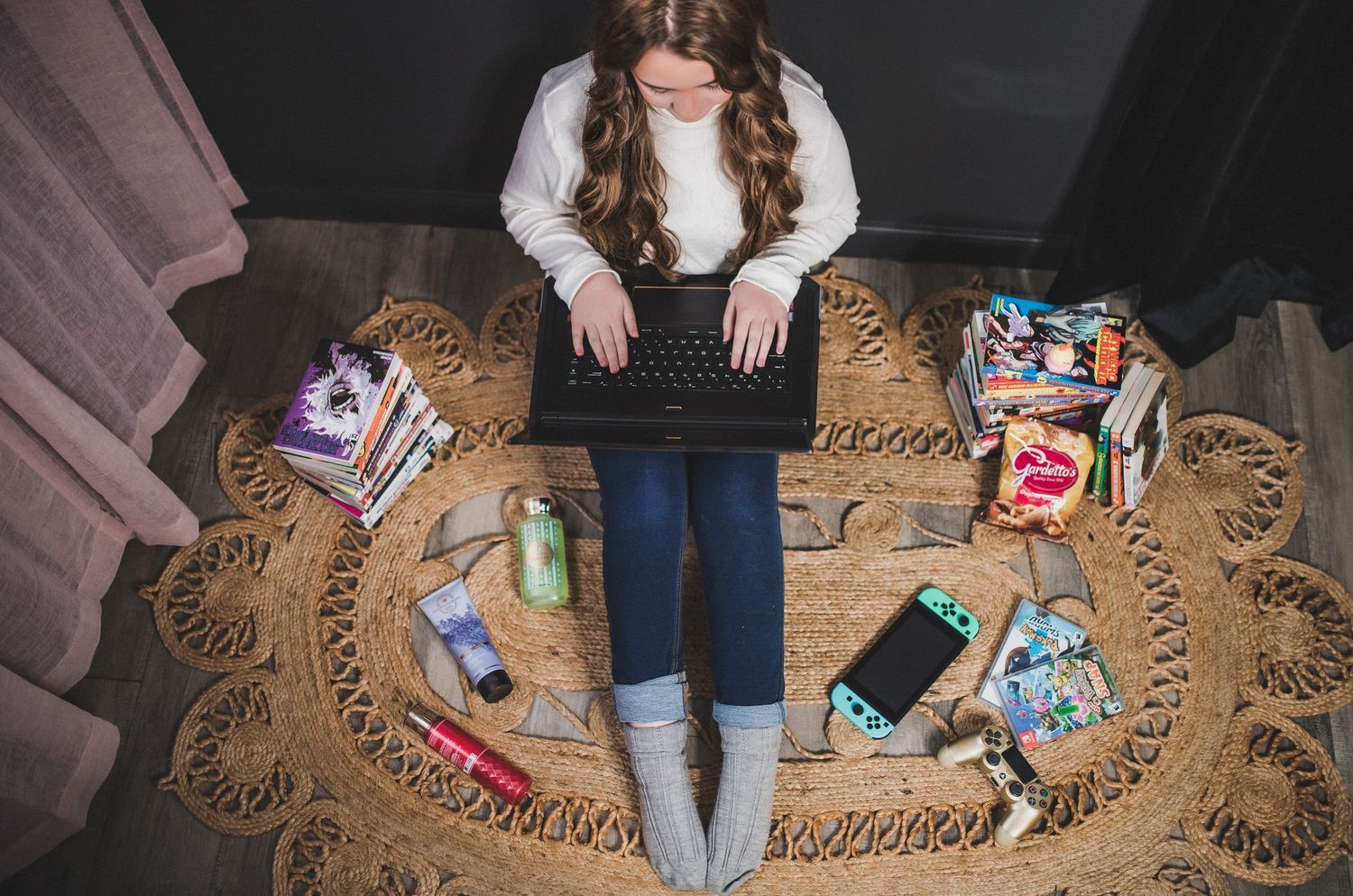 Top view of laptop user sitting on woven rug surrounded by scattered snacks, candy and a smartphone.