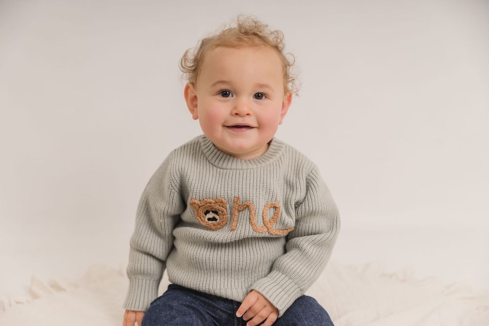 Smiling baby wearing grey sweater and jeans sits on white backdrop during photo session. Photographer. Hull. East Yorkshire. Photography