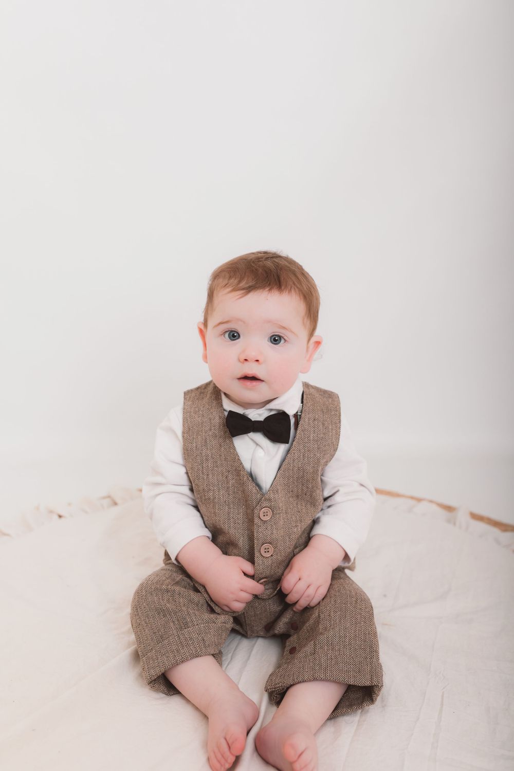 Baby in formal brown vest and bowtie outfit sitting against white backdrop.