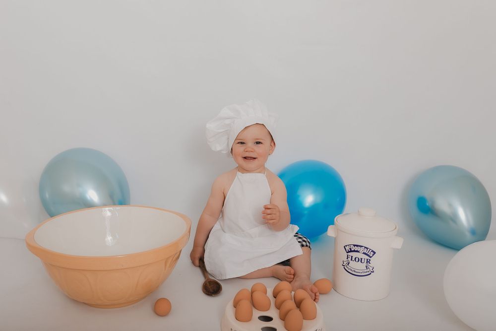 Baby in chef outfit sitting with baking props and blue balloons against white background. Photographer. Hull. East Yorkshire. Photography