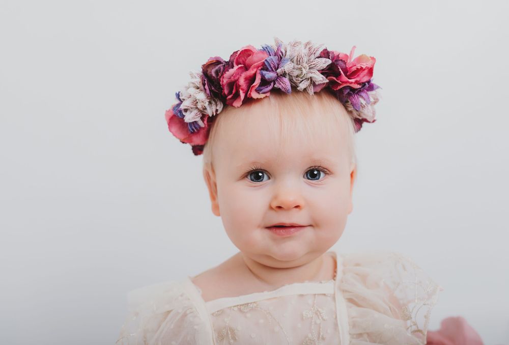 Baby wearing pink and purple floral crown and cream dress.