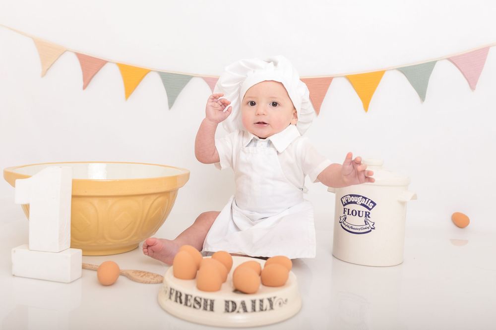 Baby in white outfit sitting among vintage milk bottles and baking props under festive bunting. Photographer. Hull. East Yorkshire. Photo