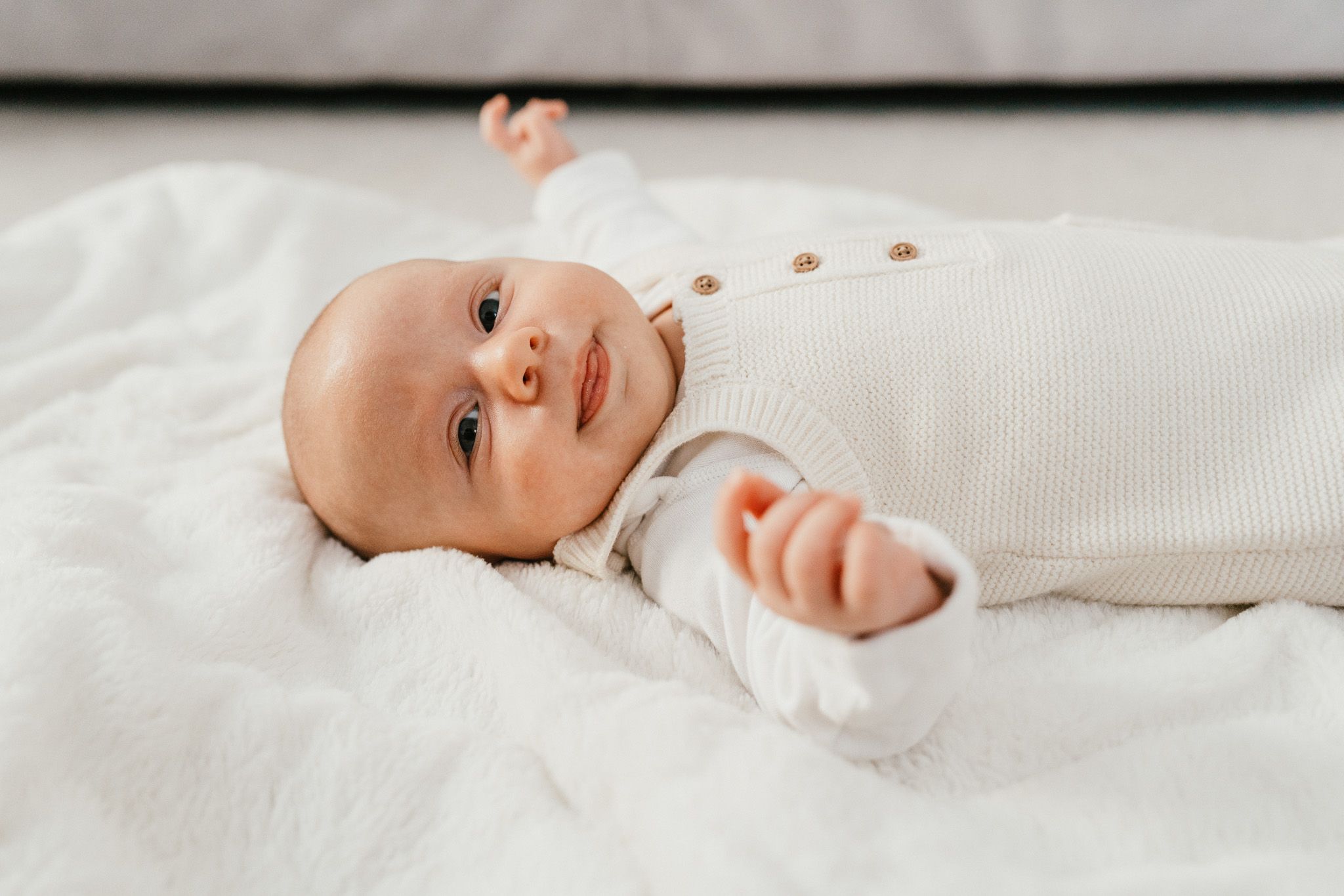 Baby in white onesie lying on white bedding looking up with bright expression.