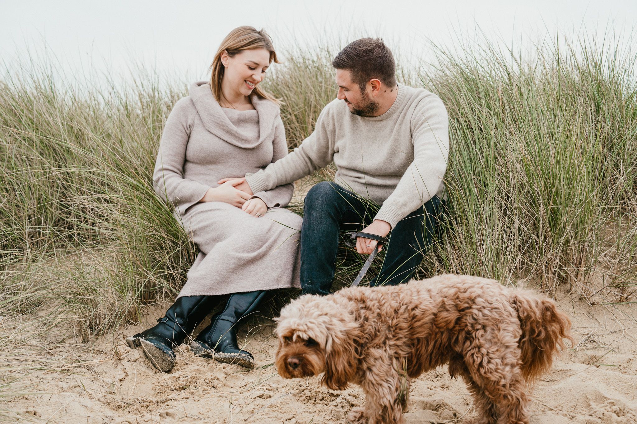Couple sitting on beach sand with brown dog among tall grass dunes.