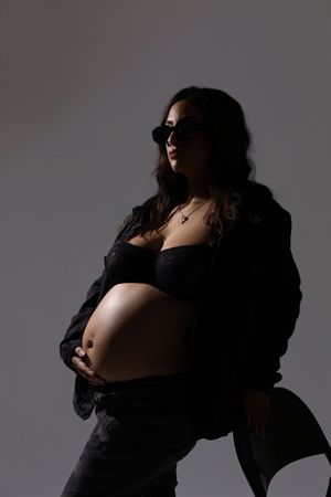 Pregnant woman in sunglasses and an open dark jacket cradles her bare belly in a moody, dramatic studio maternity portrait.