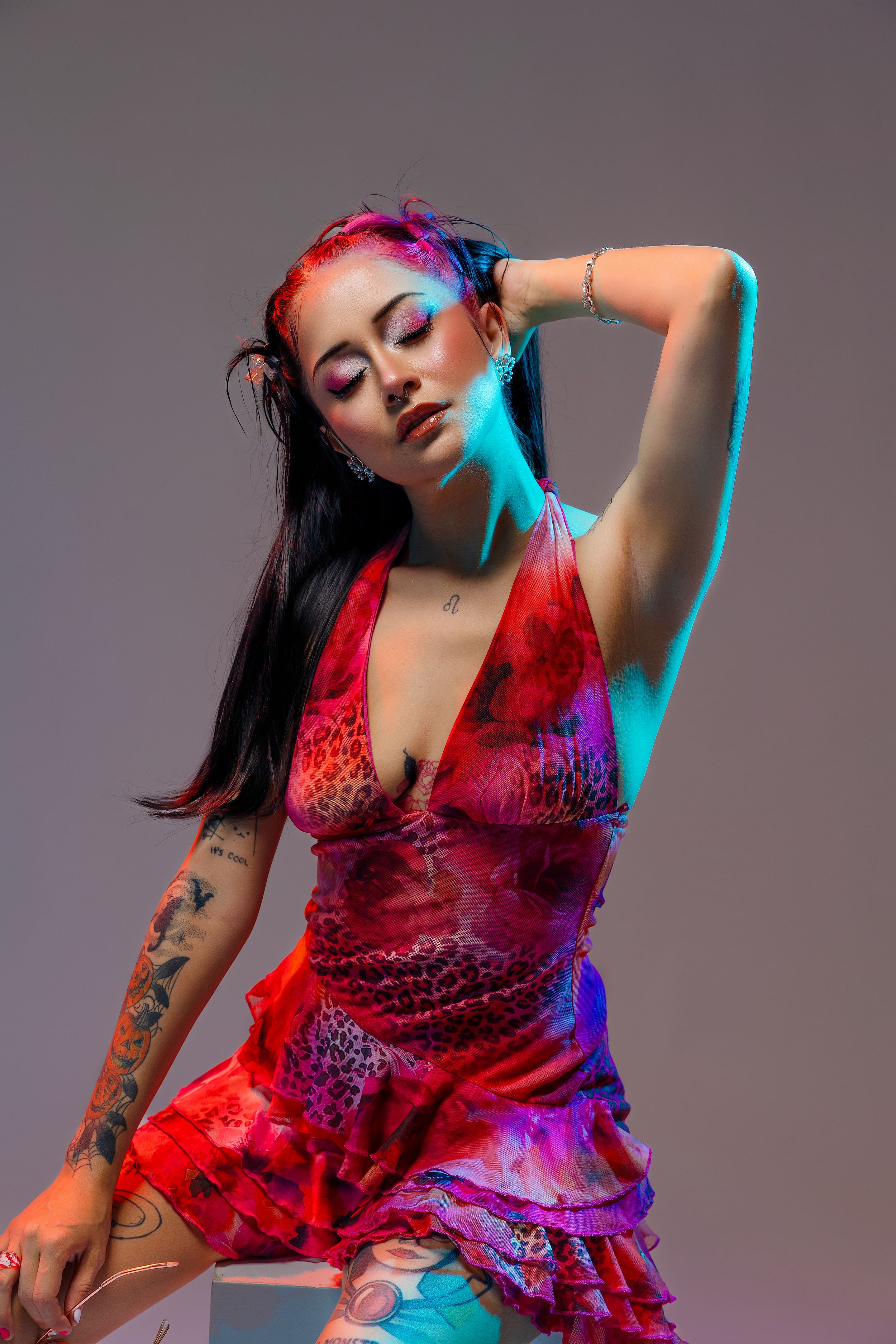Woman with long dark hair and pink highlights poses in a red patterned ruffled dress with tattoos against a gray studio backdrop.