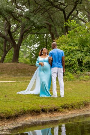 A couple in light blue and white attire stands near a pond's edge in a lush green park setting.