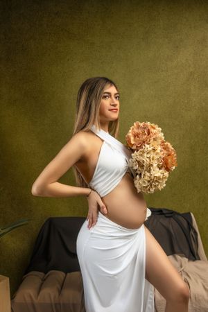 Artistic maternity portrait with flowing white dress and peach-colored hydrangea bouquet against olive textured backdrop.