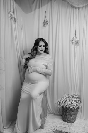 Black and white maternity photo with a flowing white gown against draped fabric backdrop and hanging dried flowers.