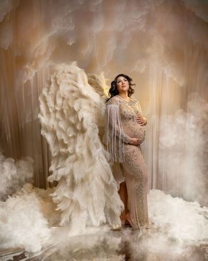 An elegant maternity photo with smoke effects, featuring a flowing white gown and angel wings in a dreamy studio setting.