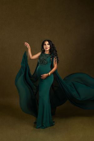 Elegant maternity photo featuring flowing emerald green gown with cape dramatically billowing against dark brown background.