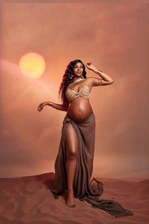 A pregnant woman in flowing brown dress poses gracefully against a sunset sky with orange sun in desert setting.