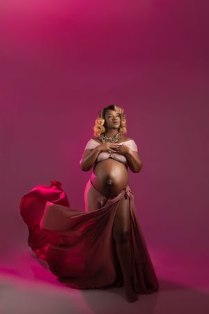 Artistic maternity photo with flowing burgundy fabric against pink backdrop in dramatic lighting.