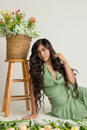 Green dress and flowing long dark hair complement a rustic scene with flowers on wooden stool and scattered petals.