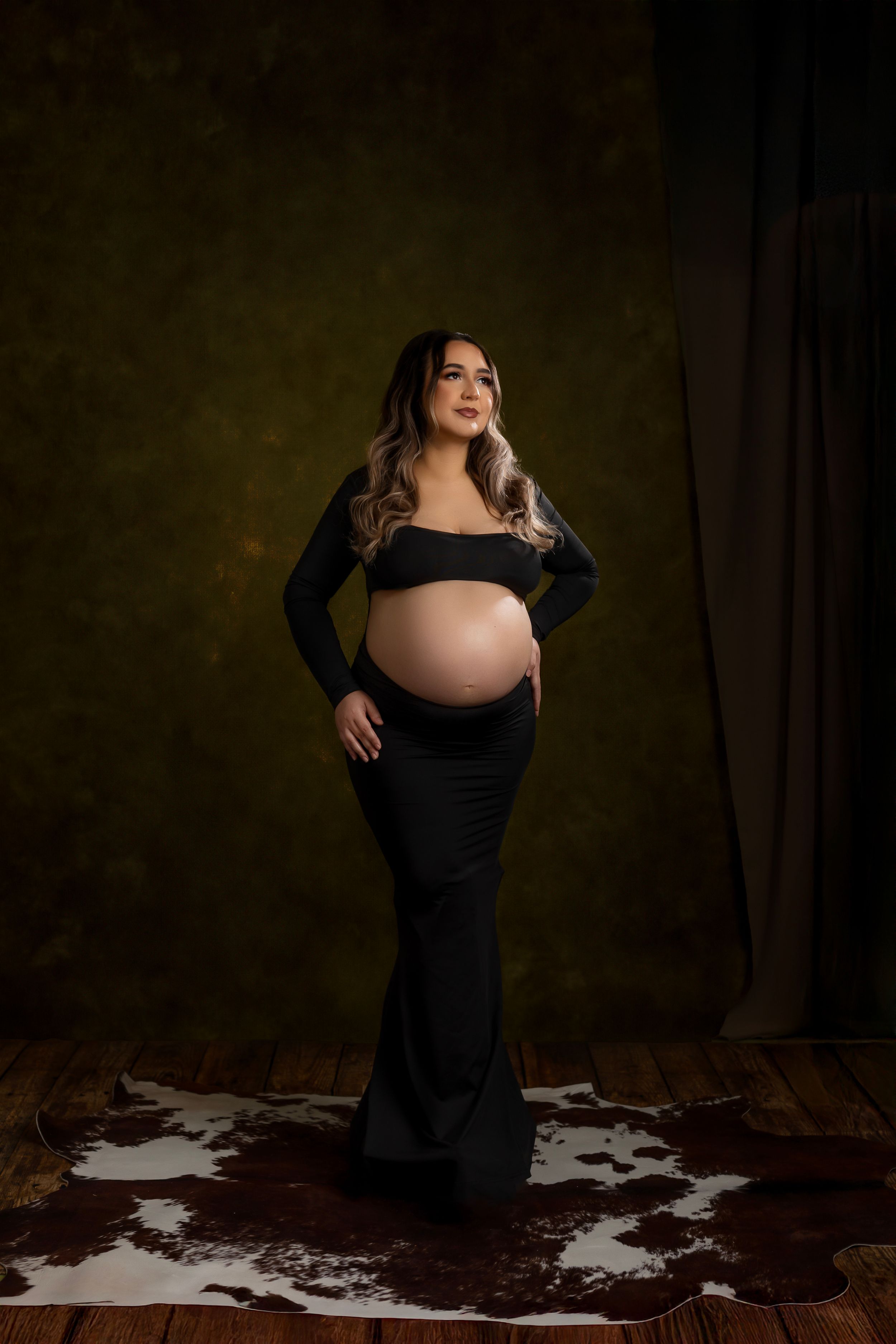 A maternity photo series showing a woman in a black dress posing against a dark backdrop with dramatic lighting.