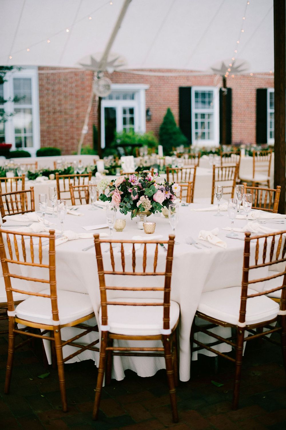 Elegant wedding reception setup with wooden chiavari chairs and white tablecloths in a brick-walled venue.