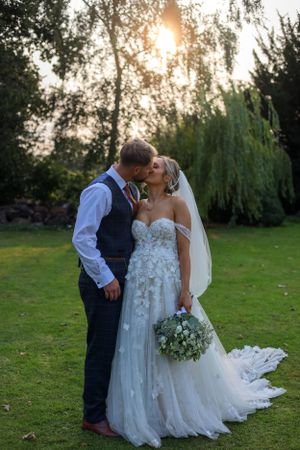 Sunset wedding portrait with couple embracing on lawn with willow trees in background.