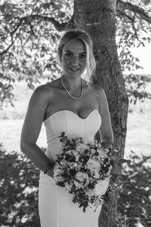 A bride in a white strapless dress holds a beautiful bouquet of mixed flowers under a tree in black and white.