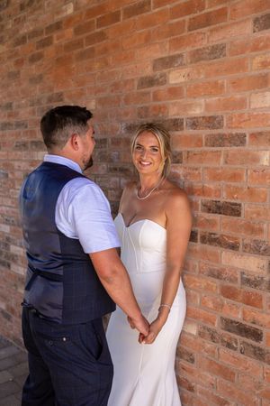 A couple in wedding attire share a candid moment against a rustic brick wall backdrop.