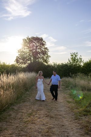 A couple walking together on a dirt path through tall grass and wildflowers at sunset.