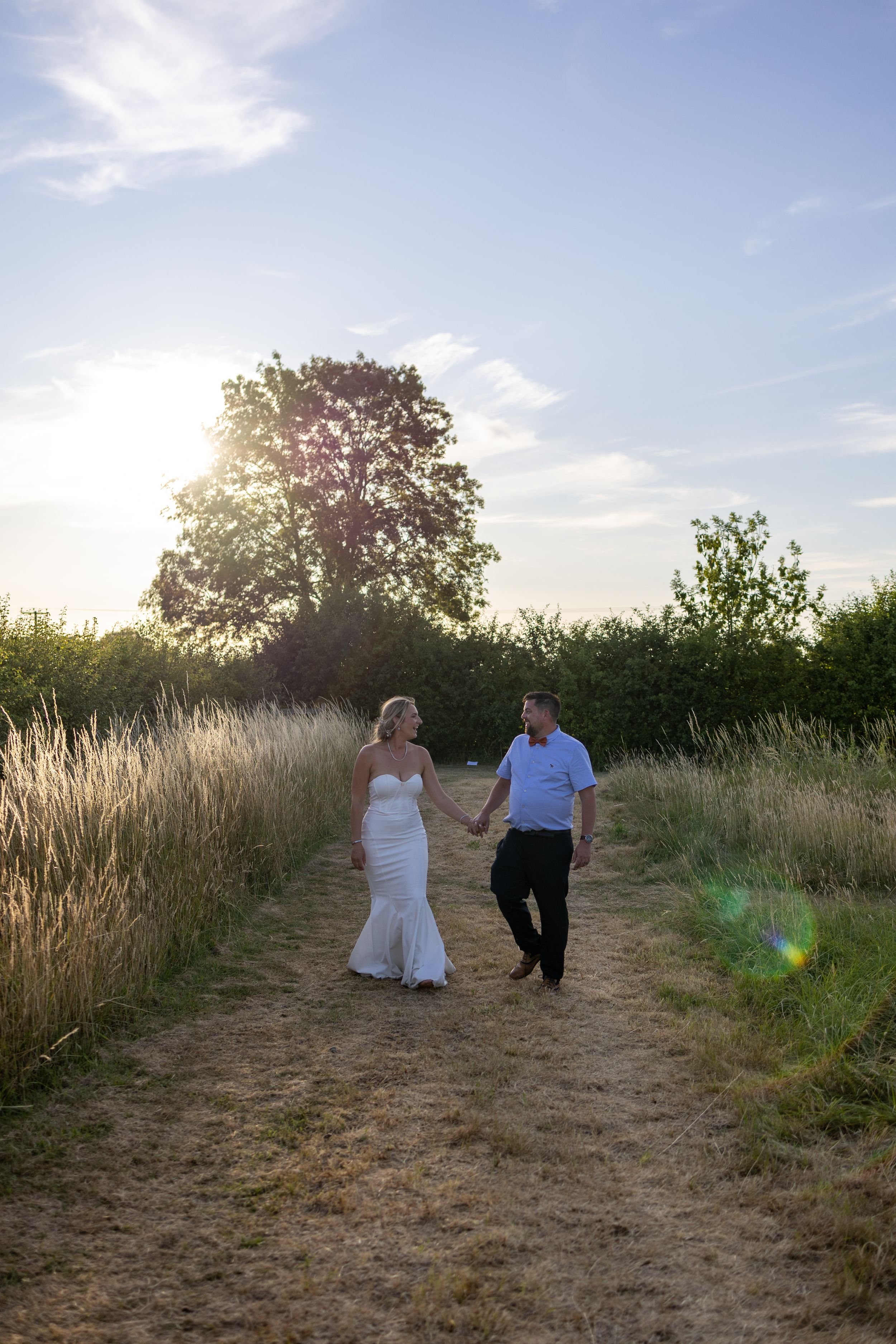A couple walking together on a dirt path through tall grass and wildflowers at sunset.