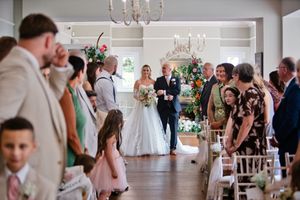Bride walking down the aisle with her father at an elegant indoor wedding ceremony surrounded by smiling guests.