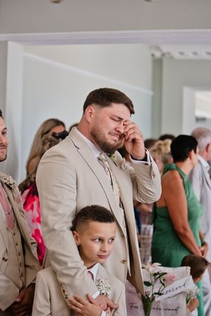 Emotional groom in beige suit wiping tears during wedding ceremony, with young ring bearer standing beside him.