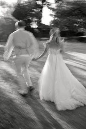 Black and white photo of bride and groom holding hands walking through garden, motion blur effect.