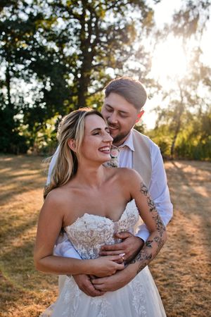 Bride and groom embrace outdoors at golden hour, she smiles in strapless lace gown as he holds her from behind.