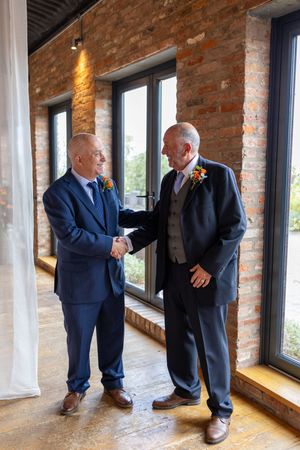 Two men in formal suits shake hands while standing in a room with exposed brick walls and large windows.