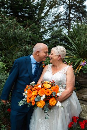 A romantic wedding couple shares tender moments with vibrant orange and white bouquet against a garden backdrop.