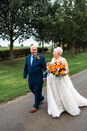 A couple in wedding attire shares a joyful moment on a tree-lined path with vibrant orange floral bouquet accents.