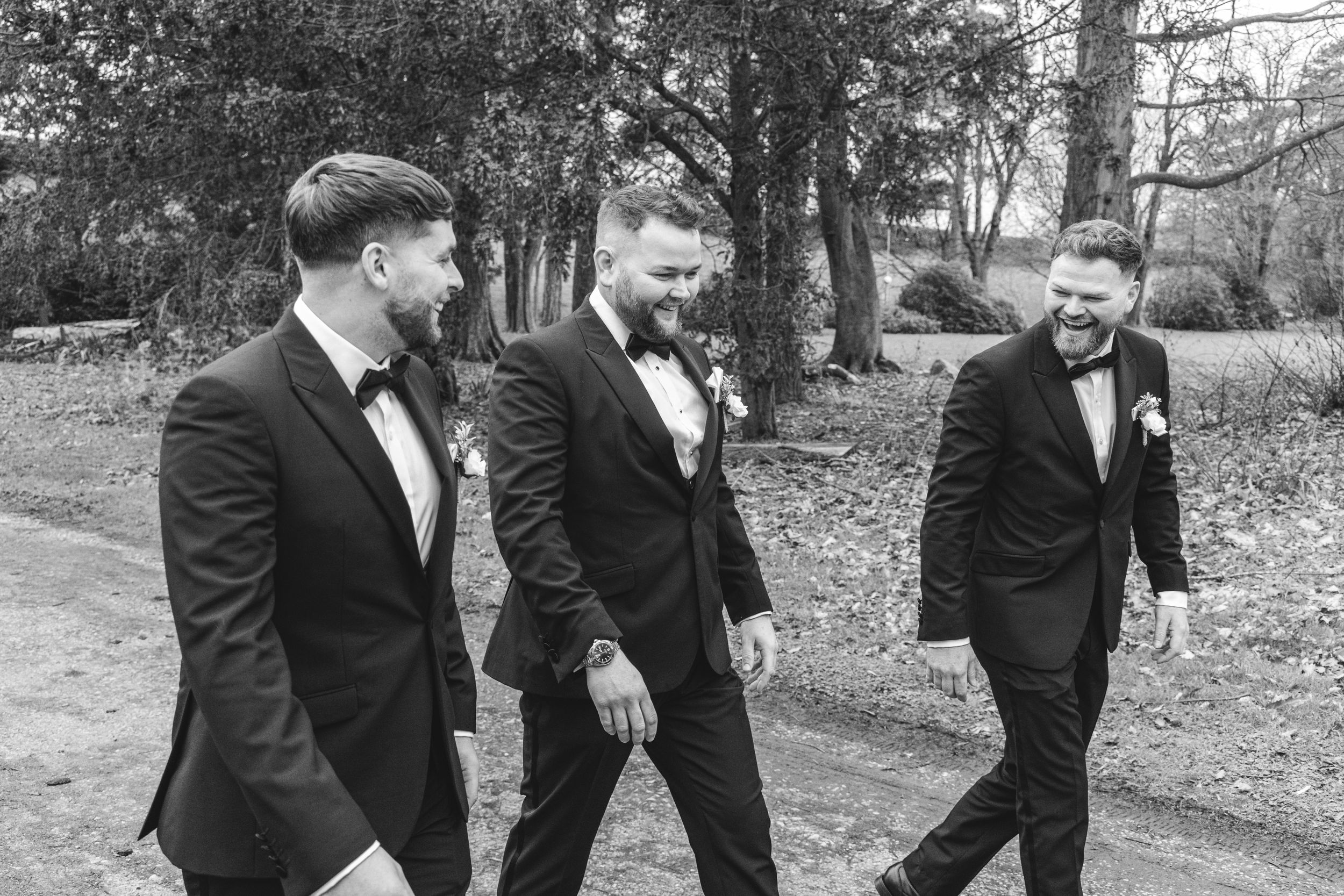 Three groomsmen in black tuxedos walk along a wooded path while sharing a laugh.
